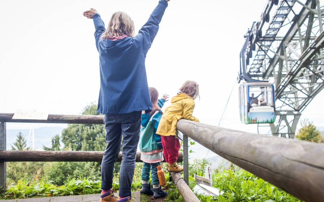 Children on a wooden railing with arms raised, watching a cable car ride in a mountainous outdoor park.