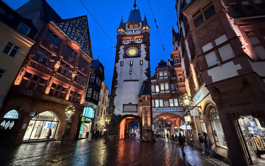 Night-time street view with a tall historic clock tower between red-brick buildings and tram tracks on a wet cobblestone road.