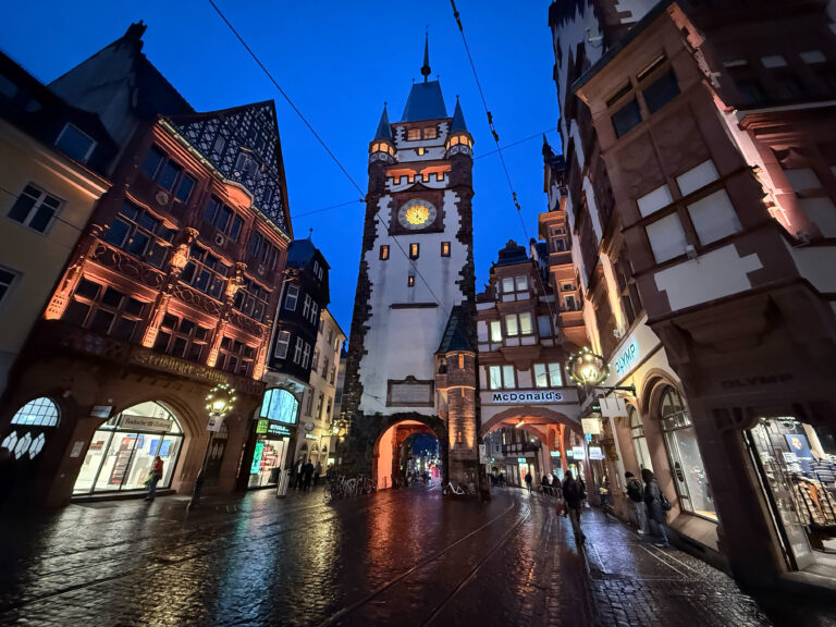 Night-time street view with a tall historic clock tower between red-brick buildings and tram tracks on a wet cobblestone road.