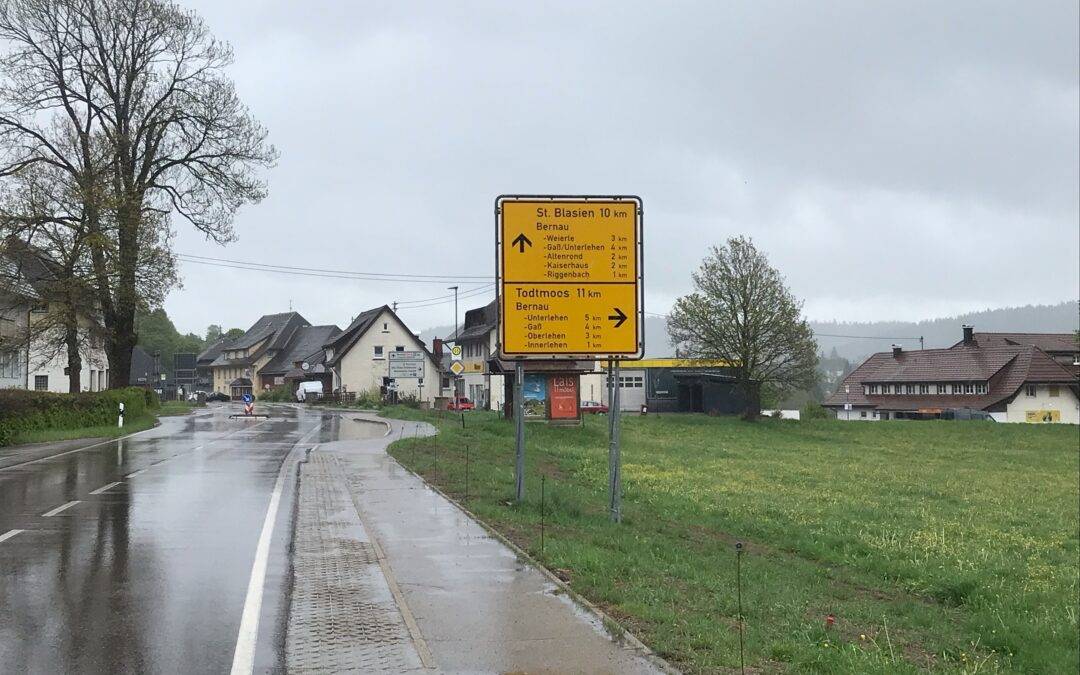 Wet street in a small town with a large yellow road sign listing nearby towns (e.g., St. Blasien, Todtmoos) under gray clouds.