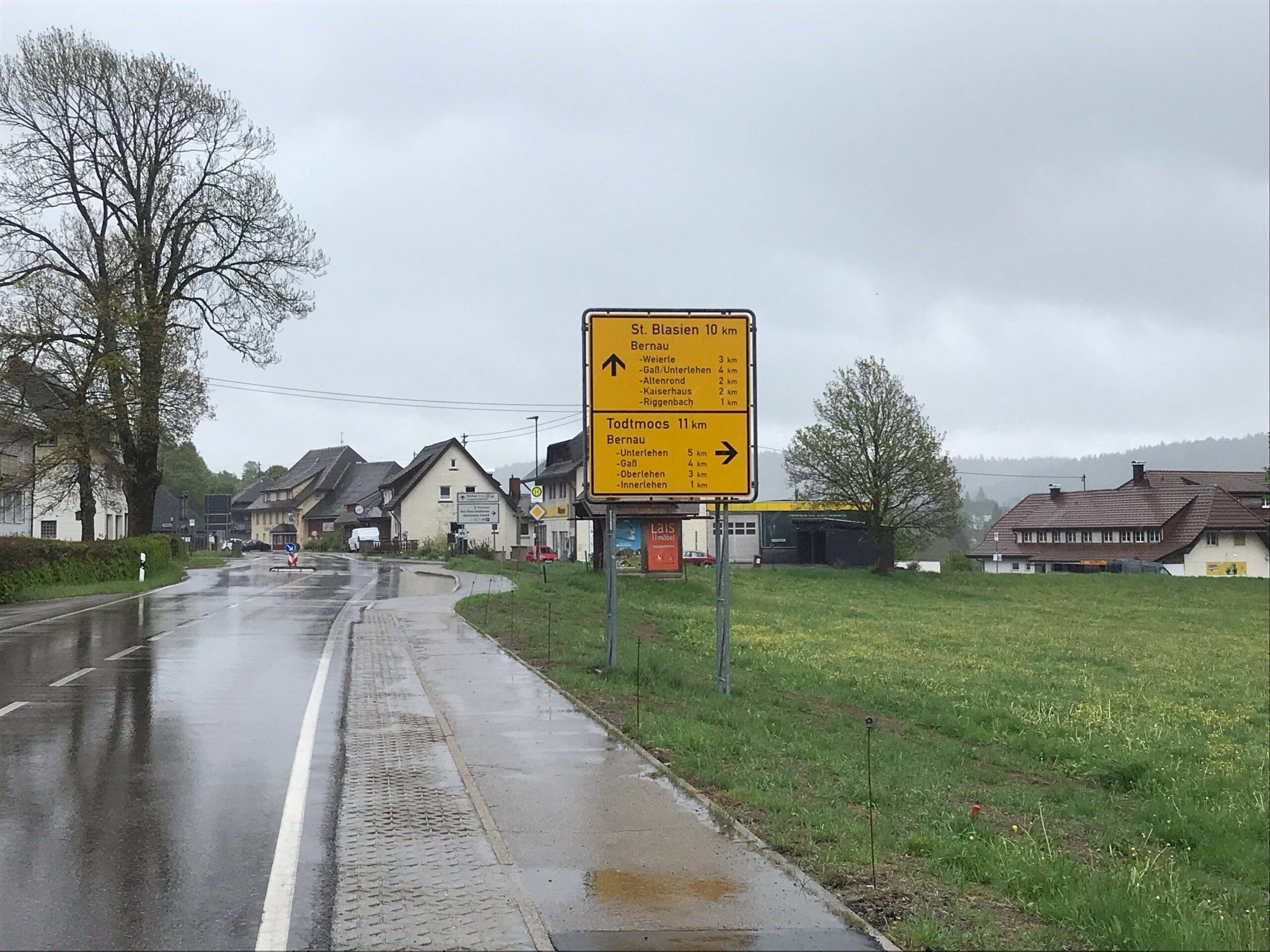 Wet street in a small town with a large yellow road sign listing nearby towns (e.g., St. Blasien, Todtmoos) under gray clouds.