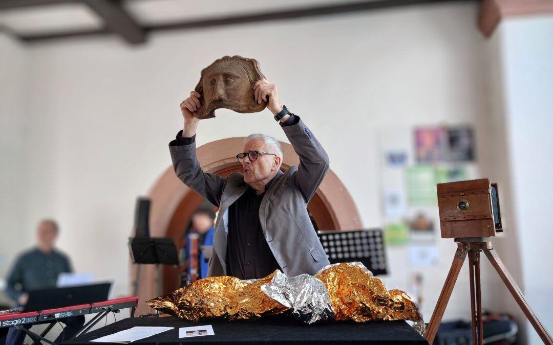 Man in a gray suit lifts a carved mask overhead as part of a performance, with foil-wrapped objects on the table nearby.