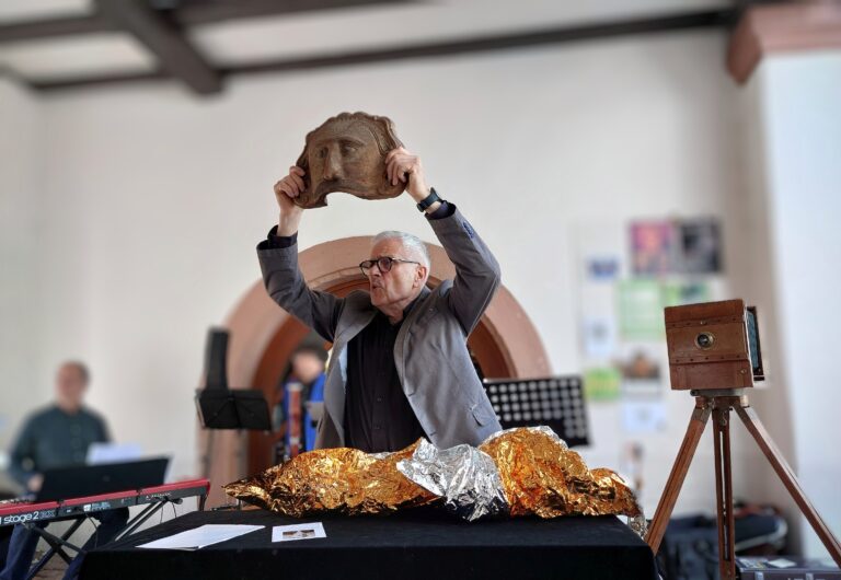Man in a gray suit lifts a carved mask overhead as part of a performance, with foil-wrapped objects on the table nearby.