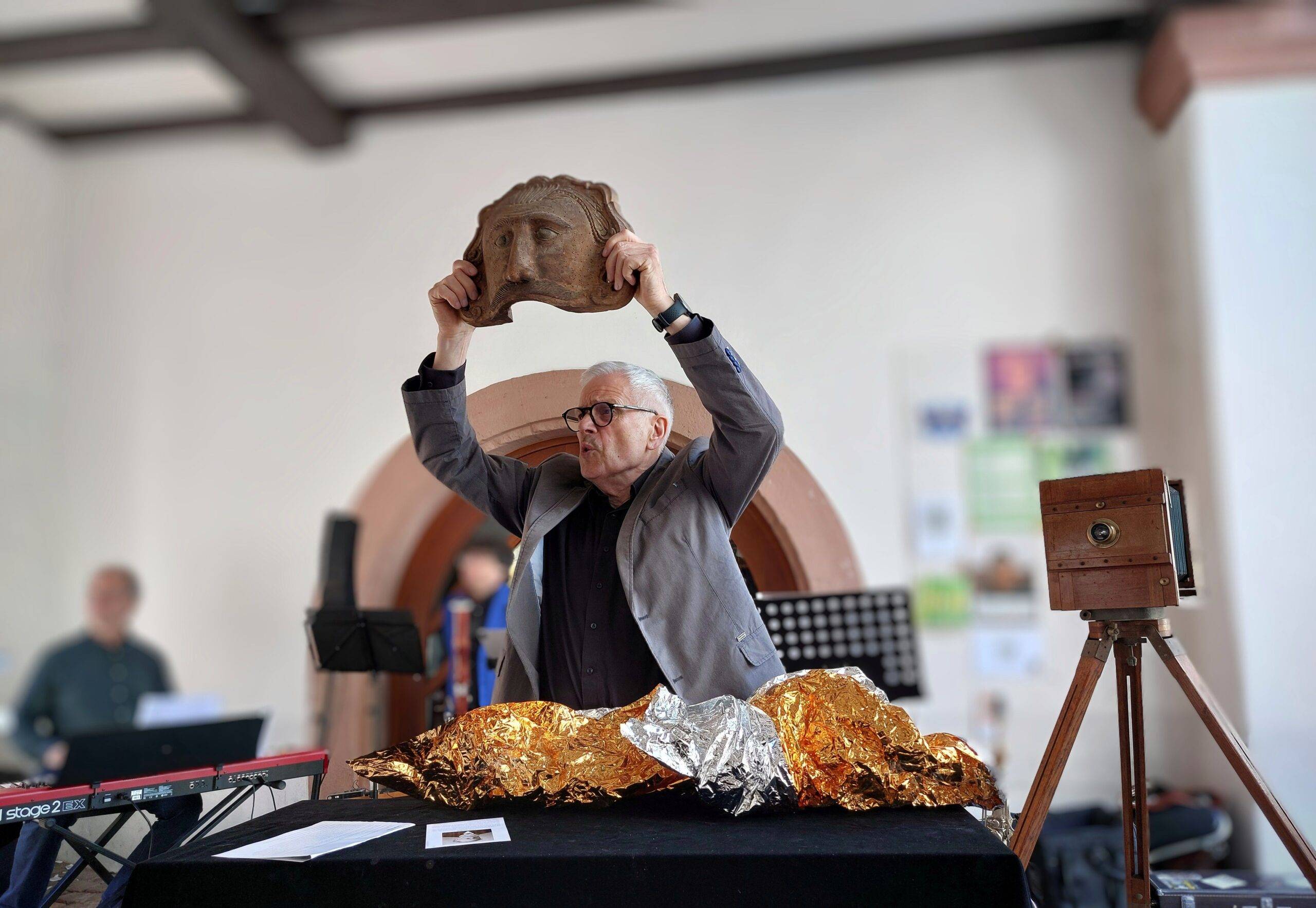 Man in a gray suit lifts a carved mask overhead as part of a performance, with foil-wrapped objects on the table nearby.