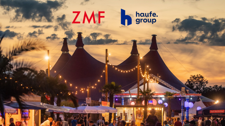 Festival scene at sunset with large tents and string lights; prominent ZMF and haufe group logos in the sky above the crowd.