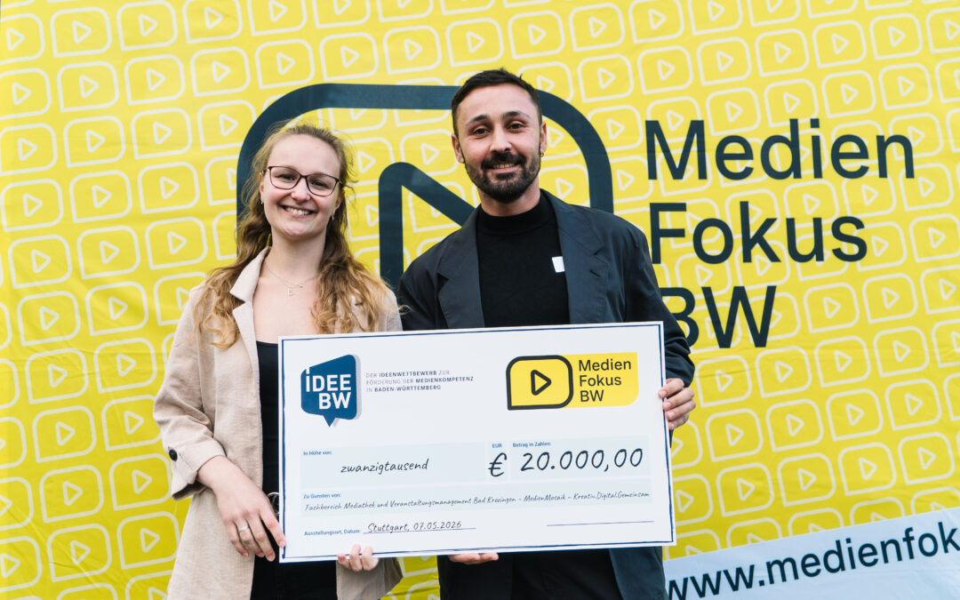 Two smiling individuals hold a large ceremonial check for €20,000 from IDEE BW to Medien Fokus BW, standing in front of a bright yellow backdrop with a repeated play-icon pattern.