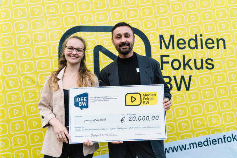 Two smiling individuals hold a large ceremonial check for €20,000 from IDEE BW to Medien Fokus BW, standing in front of a bright yellow backdrop with a repeated play-icon pattern.