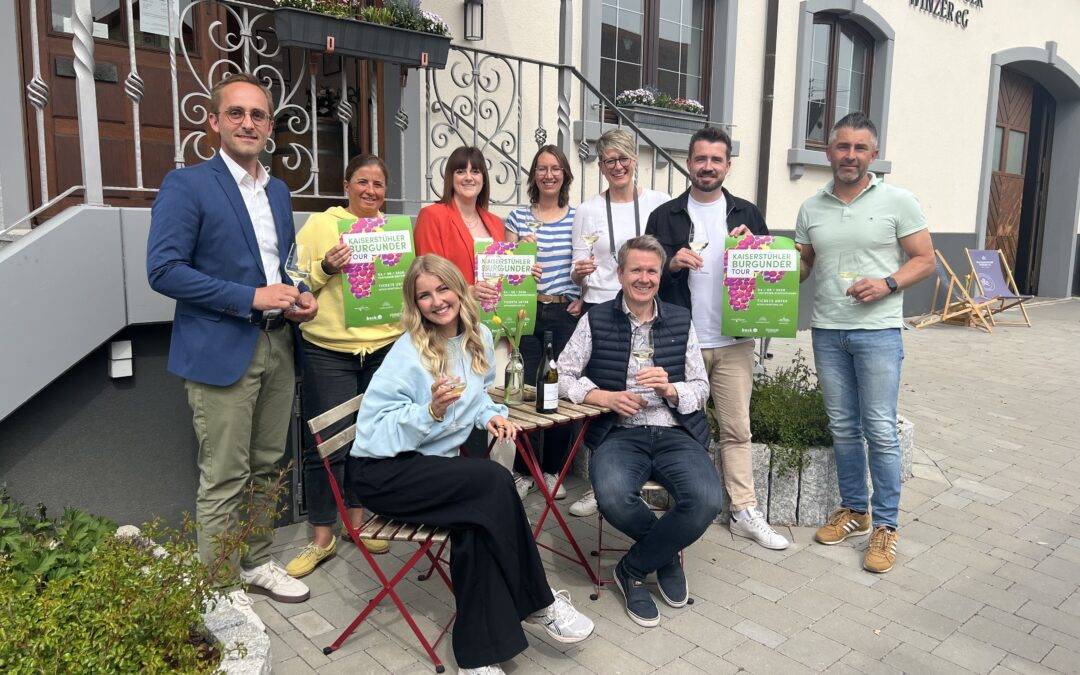 Group of eight adults posing outside a building, holding bright green event posters and wine glasses, smiling at the camera.