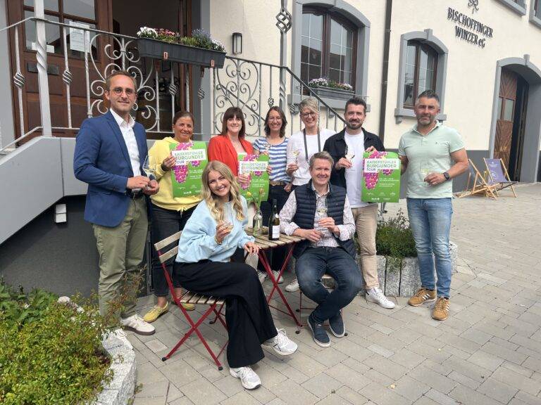 Group of eight adults posing outside a building, holding bright green event posters and wine glasses, smiling at the camera.