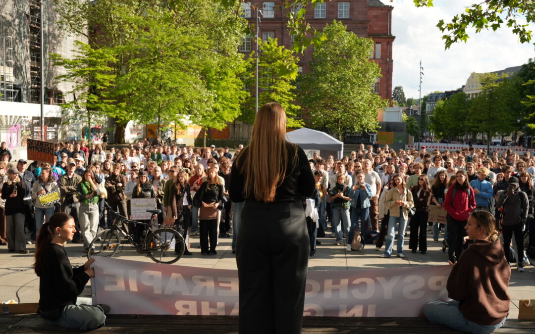 Speaker with long hair addressing a large crowd at an outdoor protest, banner in the foreground and trees and buildings behind.