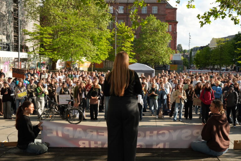 Speaker with long hair addressing a large crowd at an outdoor protest, banner in the foreground and trees and buildings behind.