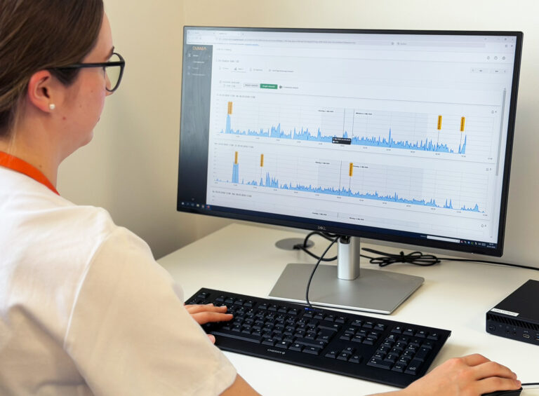 Person at a desk viewing a monitor displaying blue line charts on a dashboard, wearing glasses and a white shirt with an orange lanyard behind the keyboard.