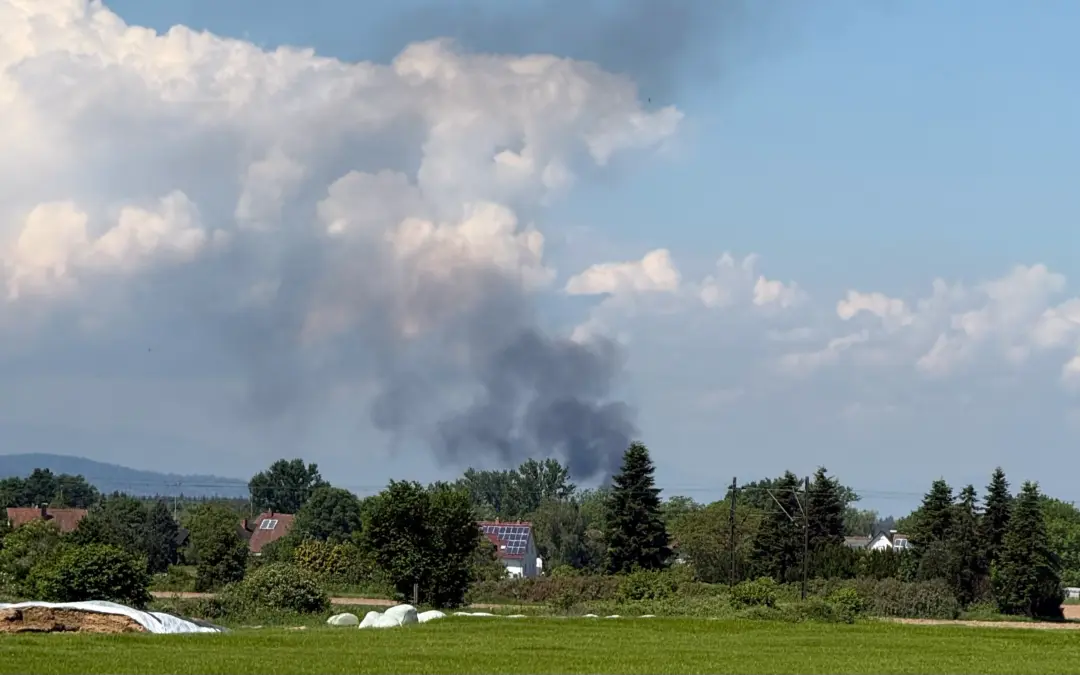 Smoke plume rising over a suburban landscape with trees and houses beneath a blue sky.
