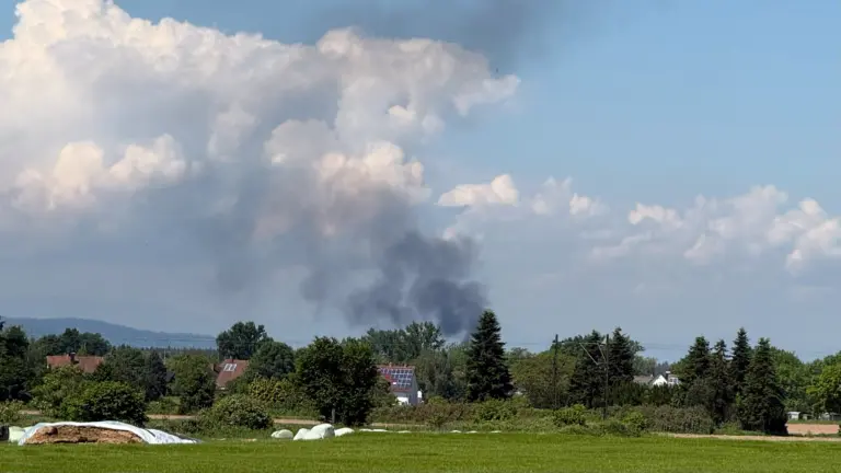 Smoke plume rising over a suburban landscape with trees and houses beneath a blue sky.