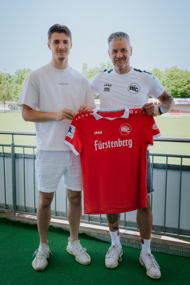 Two men stand on a balcony holding up a red soccer jersey with a BSC crest, smiling at the camera on a sunny day.