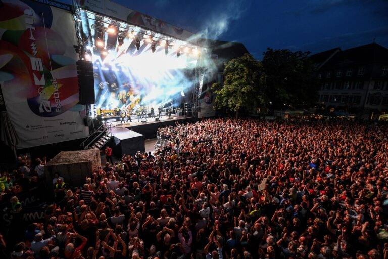 Large outdoor concert at night with a packed audience facing a brightly lit stage with performers and smoke effects on stage.