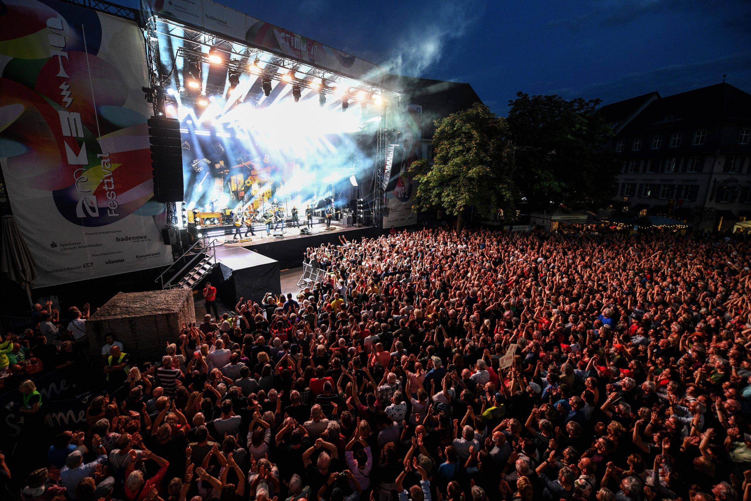 Large outdoor concert at night with a packed audience facing a brightly lit stage with performers and smoke effects on stage.