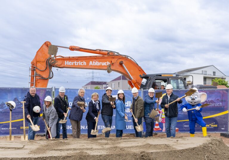 Group of people in hard hats and casual business attire using small shovels for a groundbreaking ceremony at a construction site, with an orange excavator in the background.
