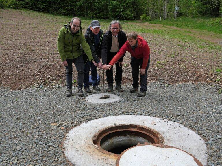 Four people outdoors around an open manhole in a gravel area, lifting the cover together.