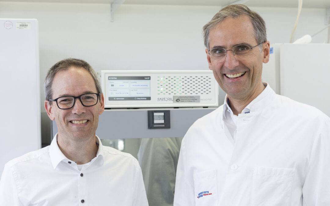 Two smiling men in white lab coats stand side by side in a laboratory with scientific equipment in the background.
