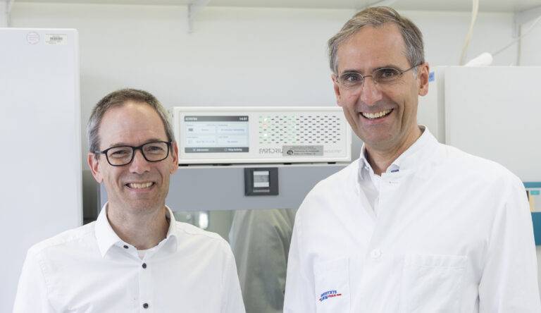 Two smiling men in white lab coats stand side by side in a laboratory with scientific equipment in the background.