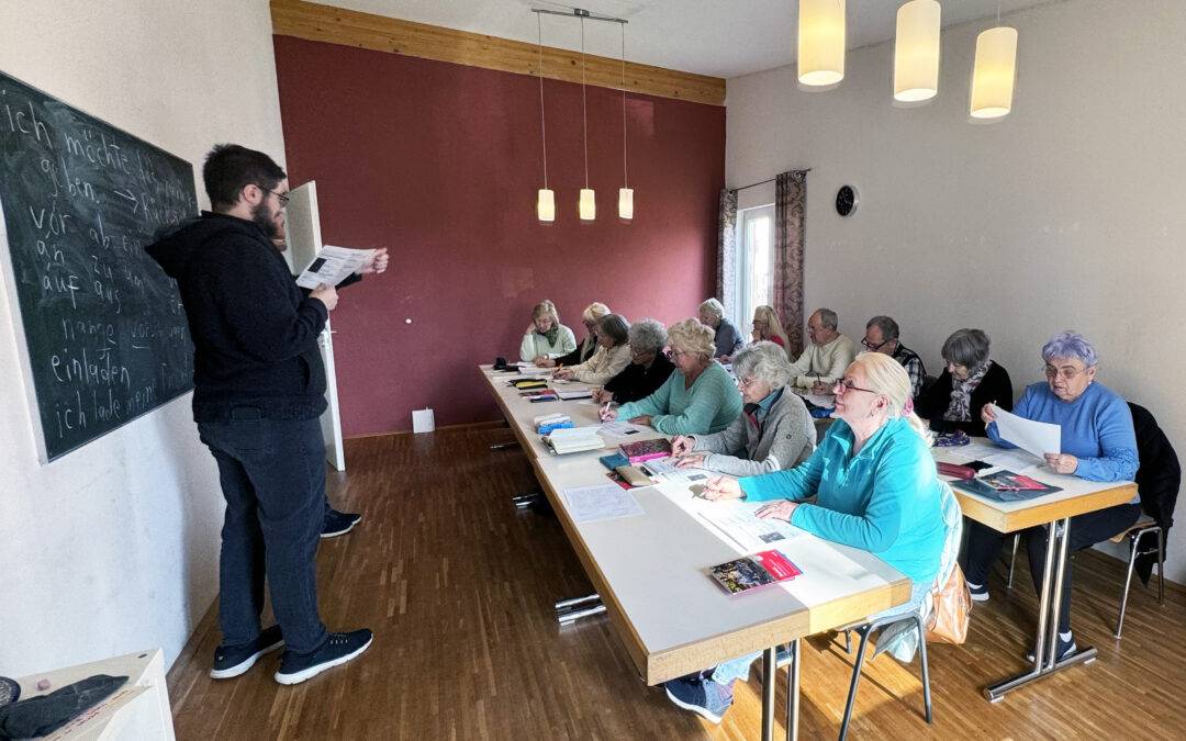 Instructor stands at a chalkboard teaching a class of seniors seated at long tables in a bright room with pendant lights. It looks like a language or writing lesson.