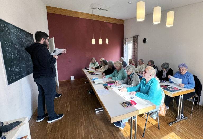 Instructor stands at a chalkboard teaching a class of seniors seated at long tables in a bright room with pendant lights. It looks like a language or writing lesson.