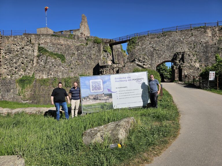 Sunlit medieval stone fortress wall with arches and a walkway; three men stand beside a large informational banner.
