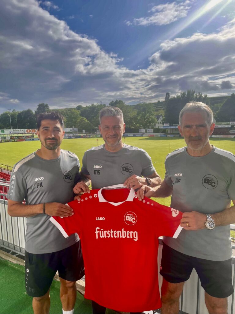 Three men in gray team shirts pose on a football field, holding a red Fürstenberg jersey with BSC logo.",