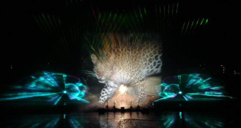 Nighttime fountain show with a leopard-print projection across a large screen and colorful laser lights over water.