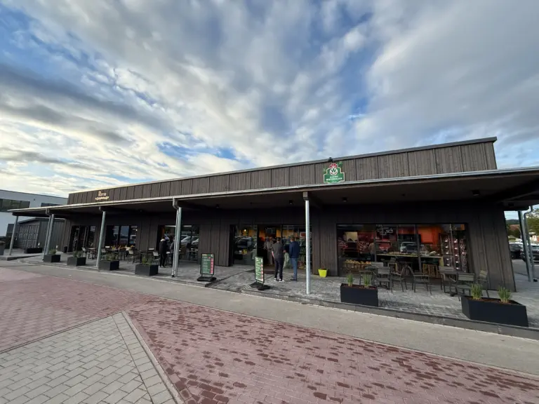 Restaurant exterior with dark wooden facade, outdoor seating, and customers under a cloudy sky.