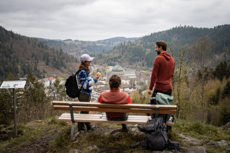 Group of hikers resting on a bench at a viewpoint, overlooking a town in a valley with forested hills and an overcast sky.