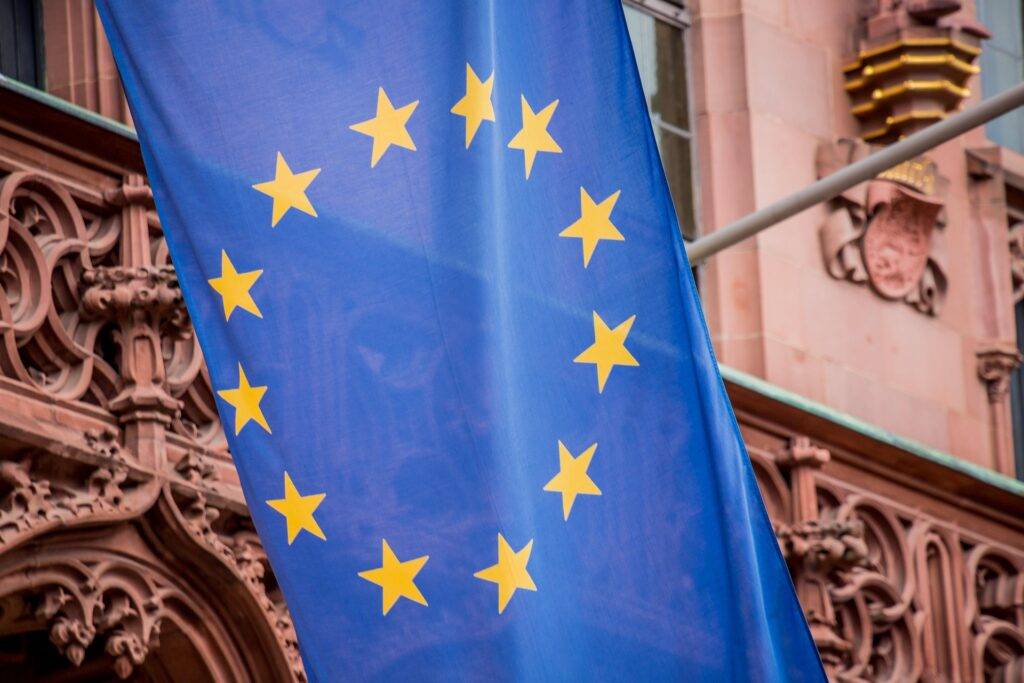 Blue European Union flag with yellow stars displayed against a historic brick facade behind it.]^(