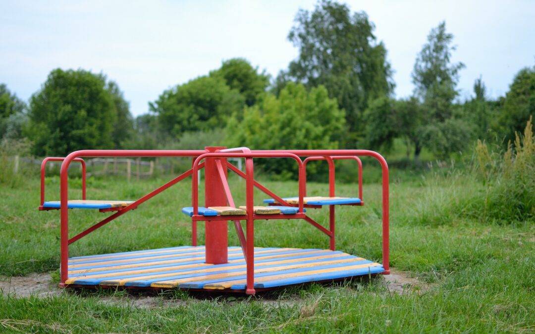 Red metal roundabout on a grassy field with a blurred treeline in the background, blue and orange slats around the platform