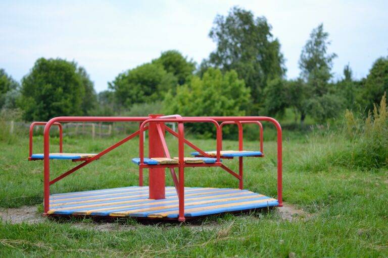 Red metal roundabout on a grassy field with a blurred treeline in the background, blue and orange slats around the platform
