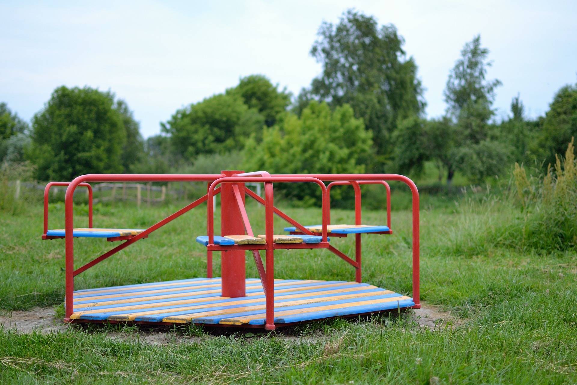 Red metal roundabout on a grassy field with a blurred treeline in the background, blue and orange slats around the platform