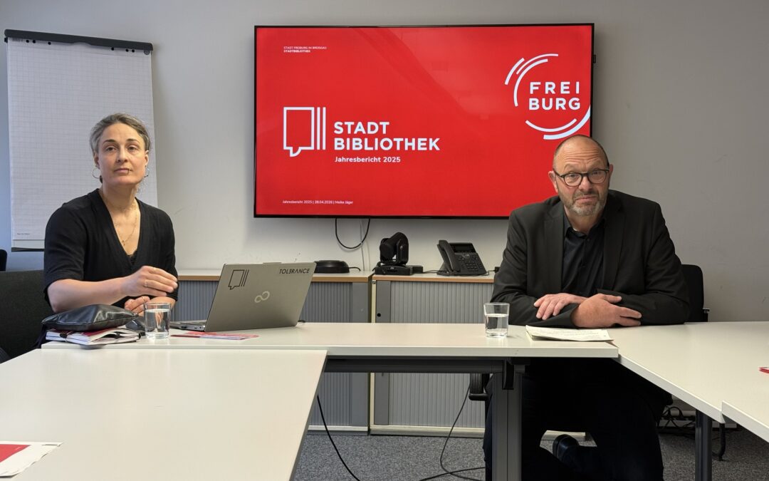 Two people sit at a conference table in a meeting room, with a large red presentation screen behind them displaying 'Stadt Bibliothek Jahresbericht 2025'.