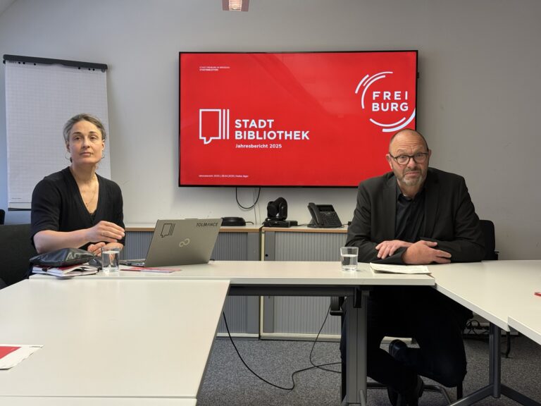 Two people sit at a conference table in a meeting room, with a large red presentation screen behind them displaying 'Stadt Bibliothek Jahresbericht 2025'.