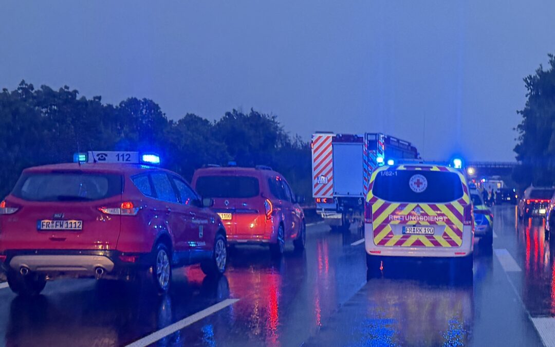 Line of emergency vehicles with blue flashing lights on a wet highway after an incident, viewed from behind a line of cars and an ambulance.