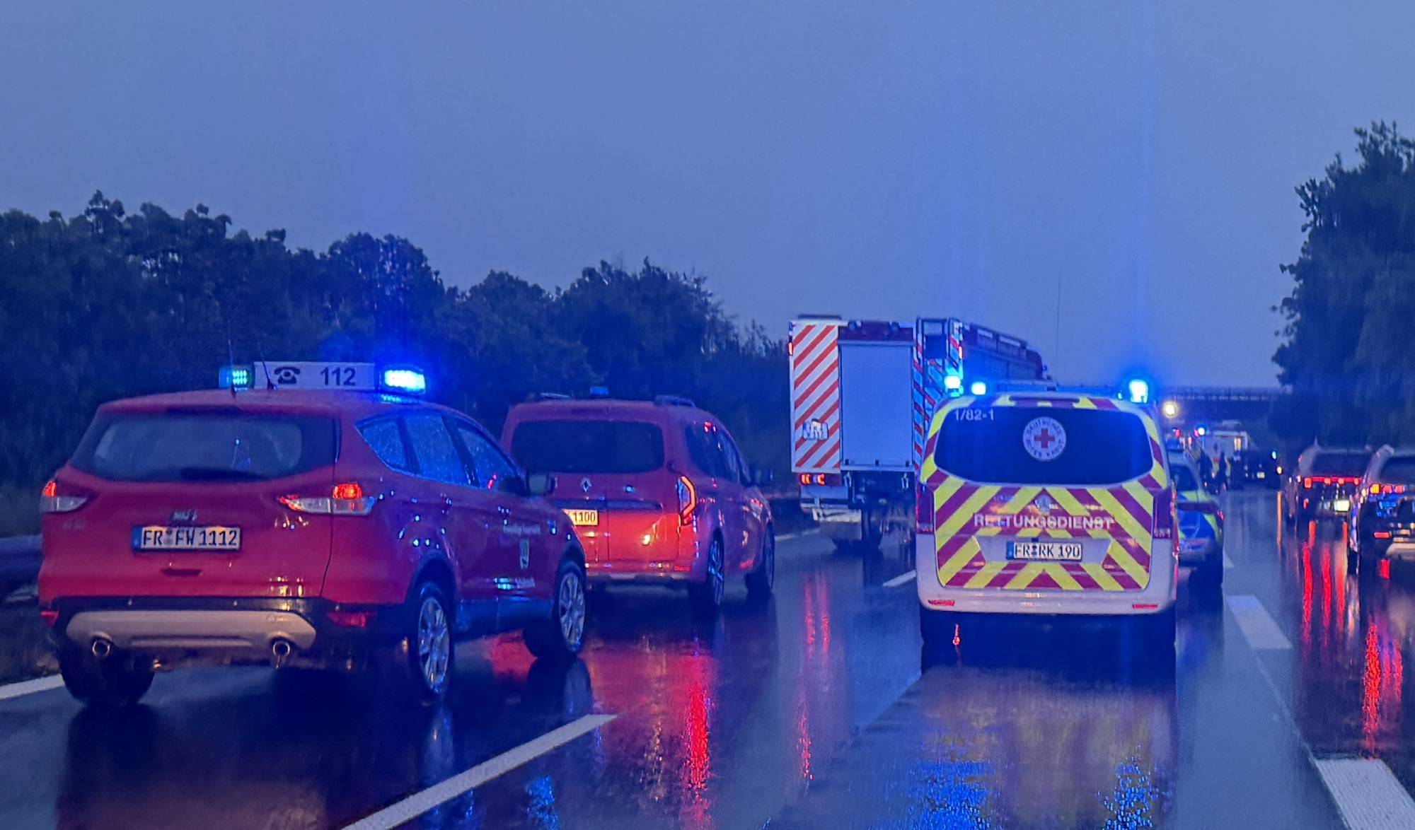 Line of emergency vehicles with blue flashing lights on a wet highway after an incident, viewed from behind a line of cars and an ambulance.