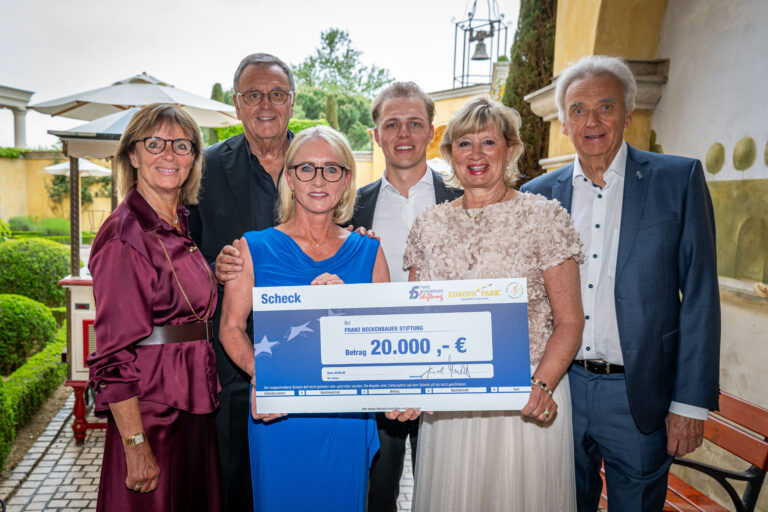 Five smiling adults stand outdoors, presenting a large ceremonial check for €20,000.