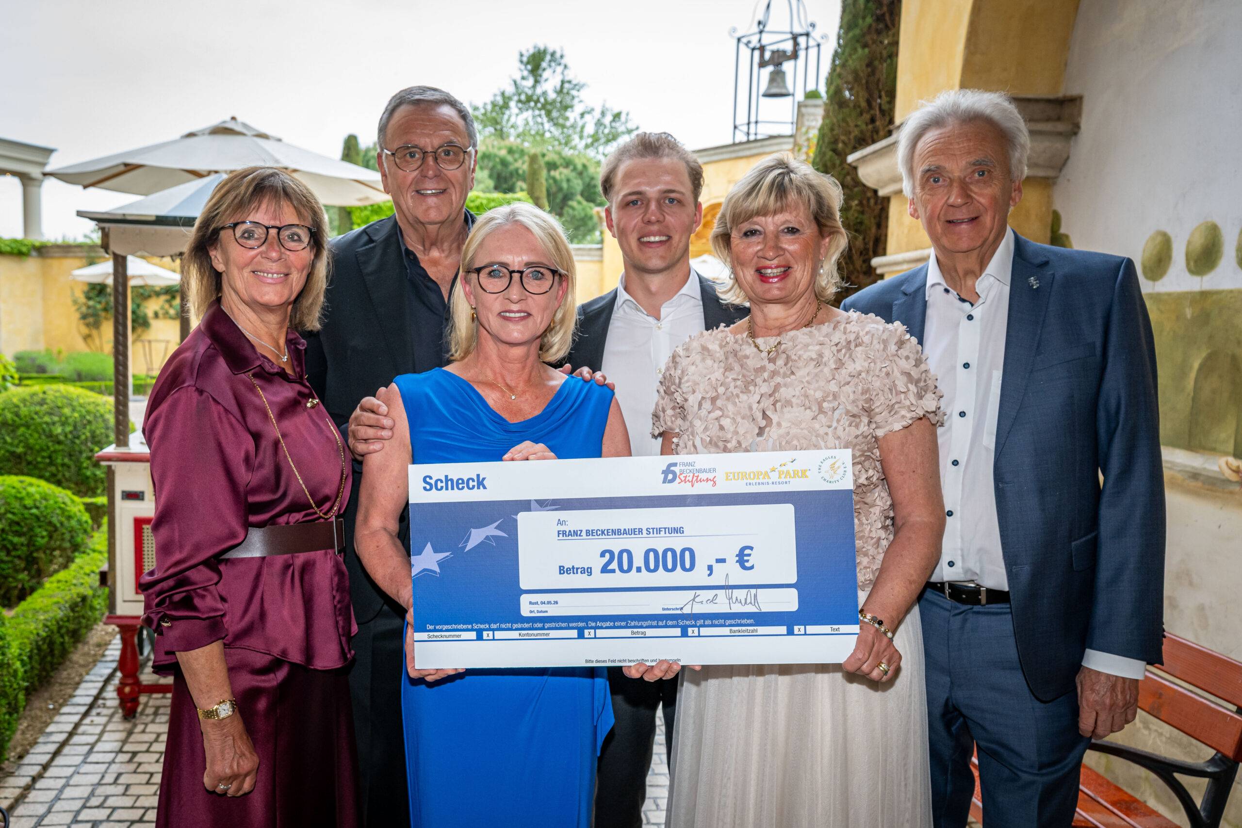 Five smiling adults stand outdoors, presenting a large ceremonial check for €20,000.