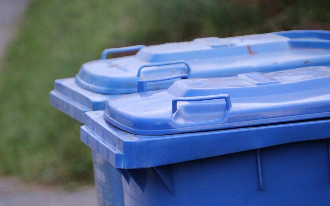 Blue plastic outdoor waste bin with lids, seen from the side outdoors on a paved area.
