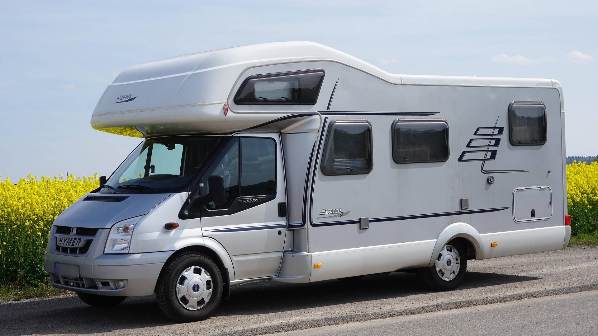 White motorhome parked on a country road beside a yellow flowering field under a blue sky.