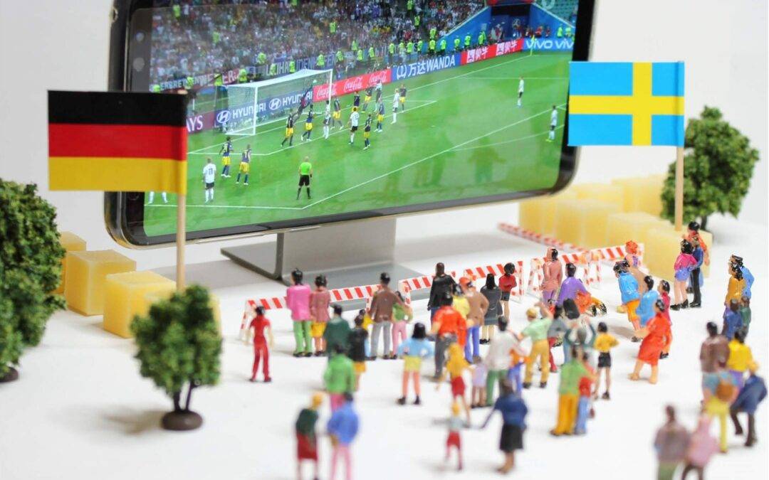 Close-up of a smartphone displaying a soccer match, with German and Swedish flags held by small figurines nearby.