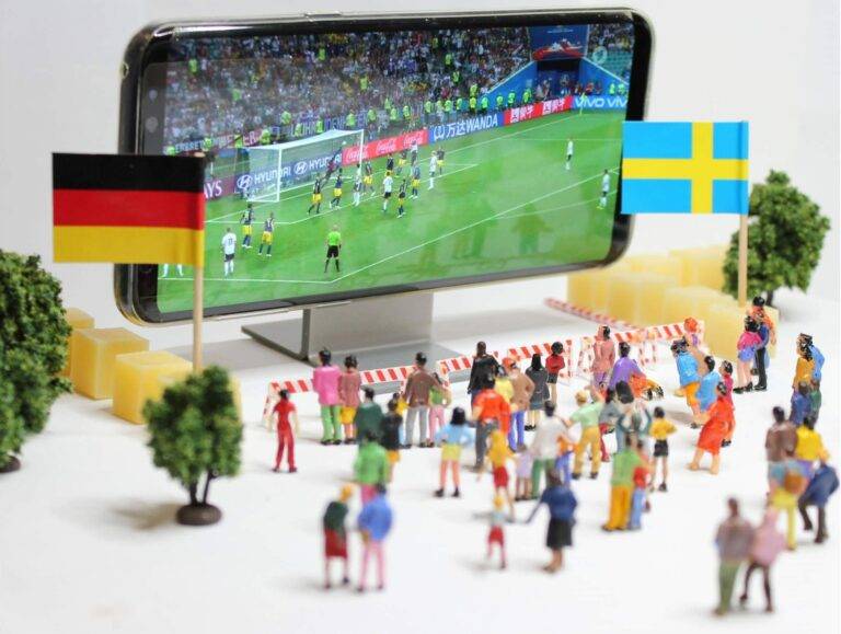 Close-up of a smartphone displaying a soccer match, with German and Swedish flags held by small figurines nearby.