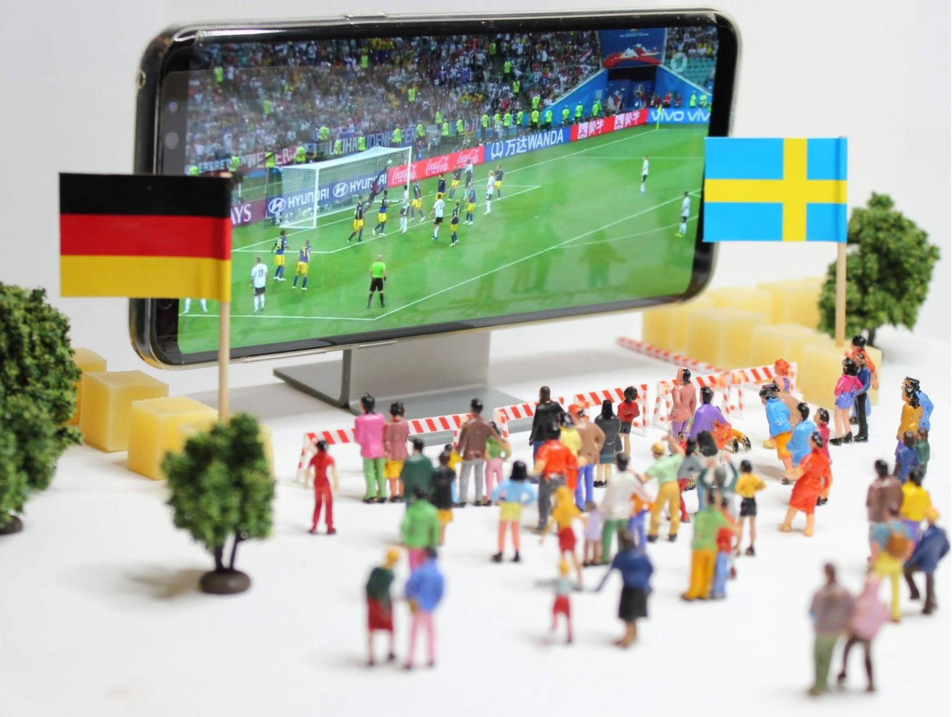 Close-up of a smartphone displaying a soccer match, with German and Swedish flags held by small figurines nearby.