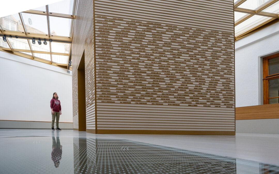 A person wearing a maroon hoodie stands in a bright museum gallery beside a tall wall covered with horizontal wooden slats bearing many small names, with a reflective floor.