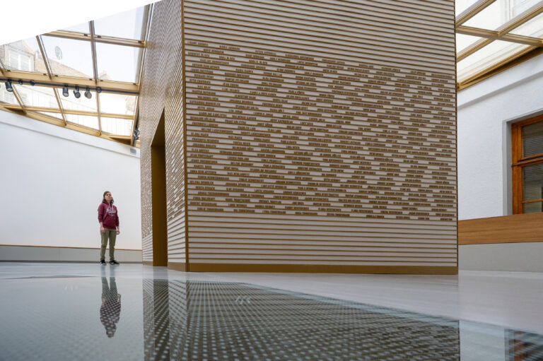 A person wearing a maroon hoodie stands in a bright museum gallery beside a tall wall covered with horizontal wooden slats bearing many small names, with a reflective floor.