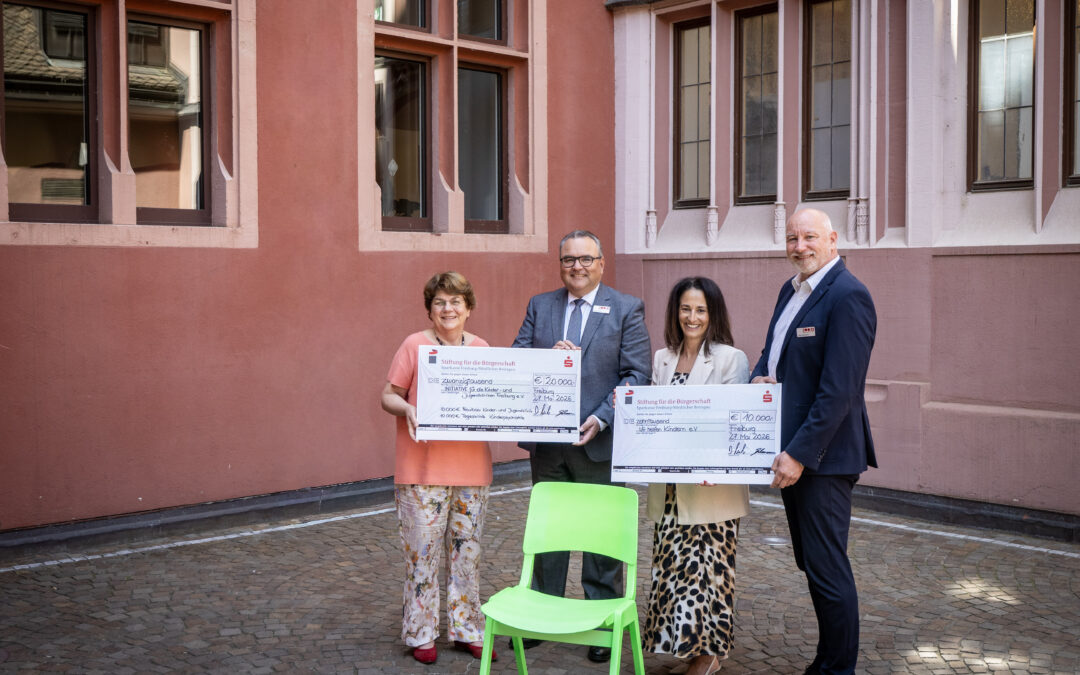 Group of four adults in smart clothing posing outdoors, holding oversized ceremonial checks in front of a pink building; a bright green chair is in the foreground.